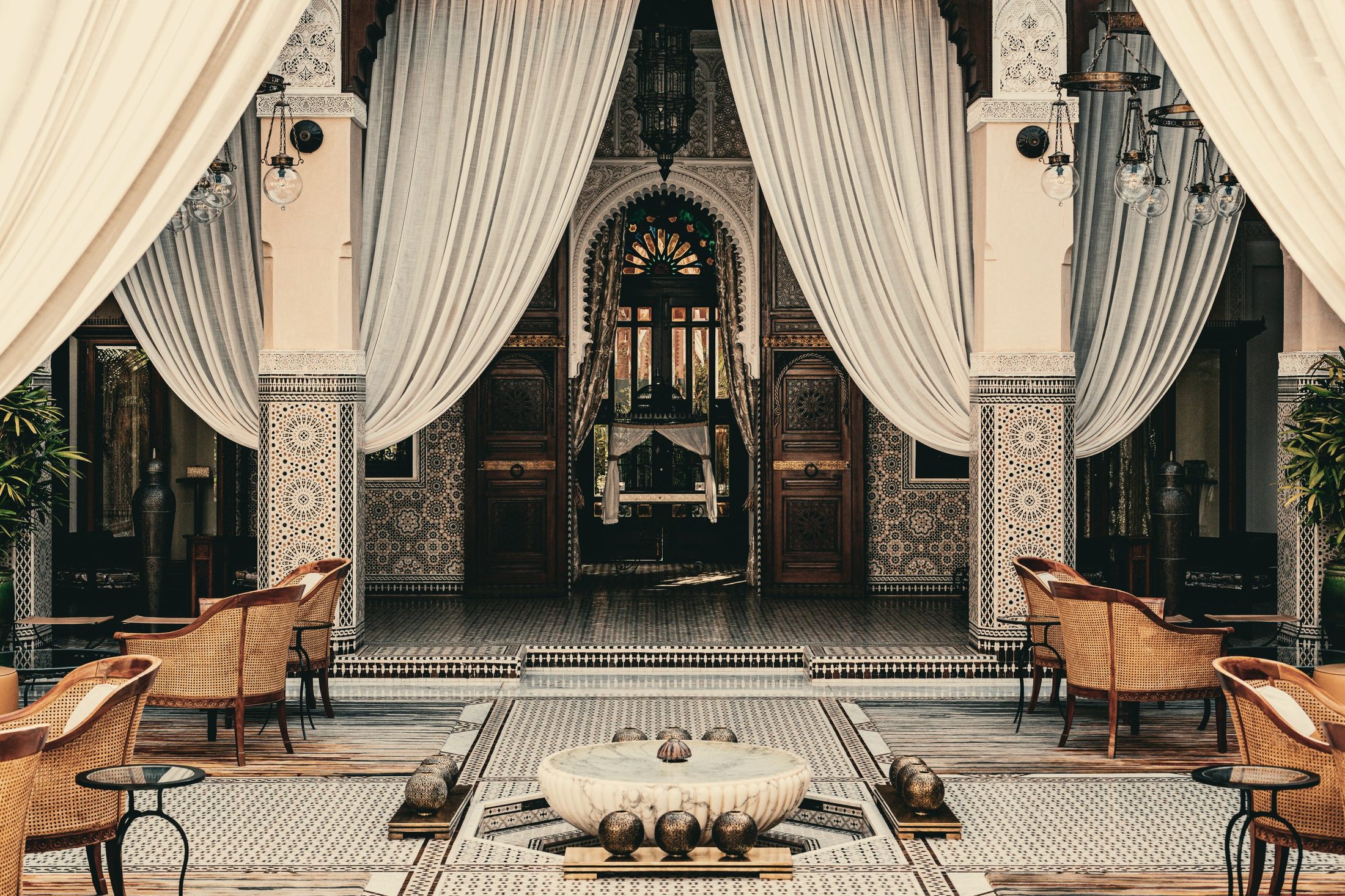 Patio d'un riad de Marrakech : colonnes de zellige, tentures claires, porte sculptée et vasque de marbre.